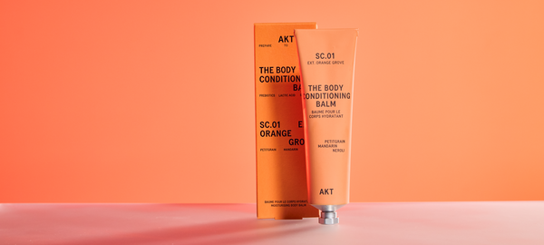 A tube of AKT Body Conditioning Balm SC.01 in Orange Grove scent, standing in front of its orange packaging box against a matching orange background. 