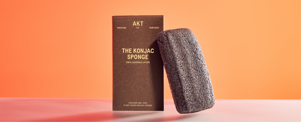 A black AKT Konjac Sponge placed beside its brown packaging box, labeled 'The Konjac Sponge' for a luxurious lather, against an orange background. 