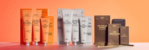 A collection of AKT body care products, including tubes of Body Wash Concentrate, Body Conditioning Balm, Deodorant Balm, The Konjac Sponge, The Applicator, and The Assistant, all displayed against an orange background.